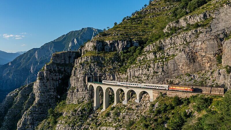 Tren sobre un puente en un paisaje montañoso, vista panorámica de acantilados y vegetación alrededor.