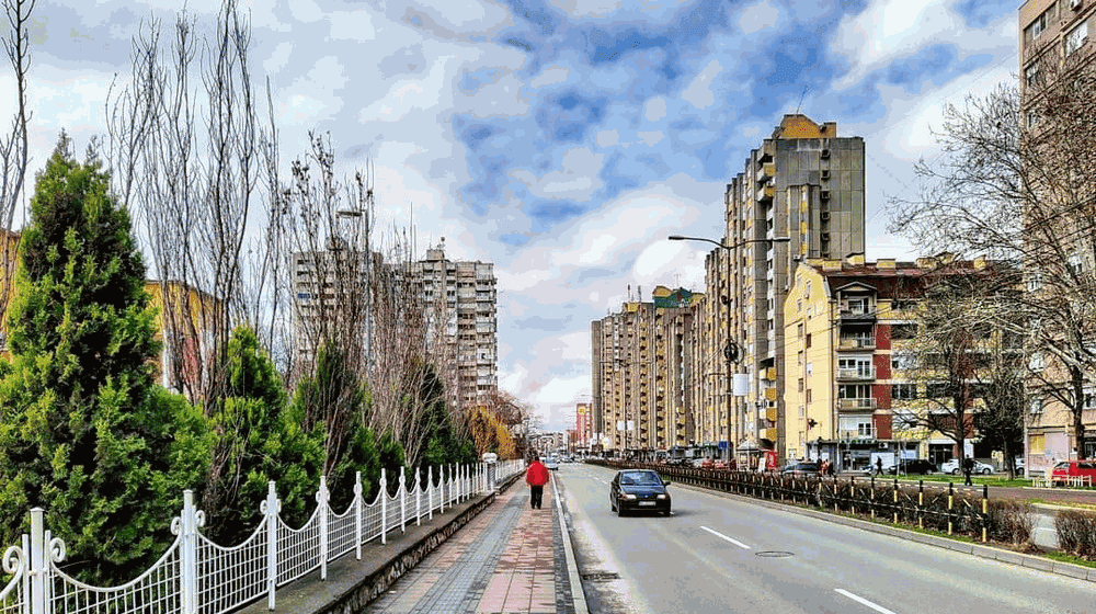 Городской пейзаж с высотными зданиями, дорогой и пешеходами в городе. Ясное небо, деревья вдоль тротуара.