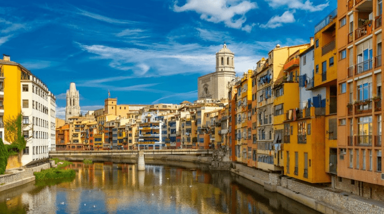 Billede med tekst: Sammenligning af ejendomspriser i forskellige regioner i Spanien på baggrund af et malerisk bybillede.