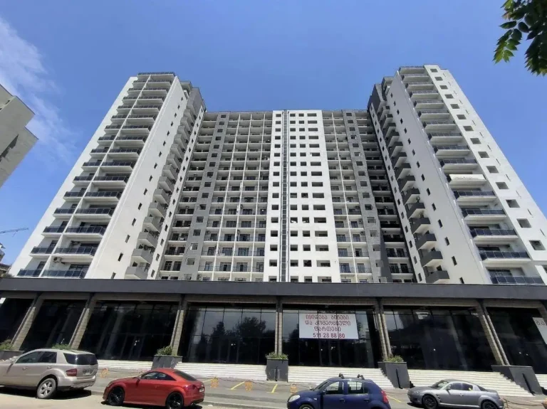 High-rise residential building against blue sky, view from below. Cars parked in the foreground.