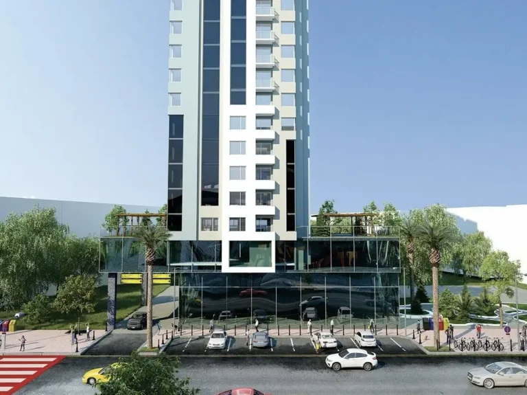 A high-rise building with a modern facade and parking lot, surrounded by greenery and palm trees, against a clear sky.