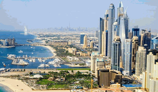 Panorama Dubaje s výškovými budovami, pobřežím a Burdž Chalífou na obzoru, slunečný den.