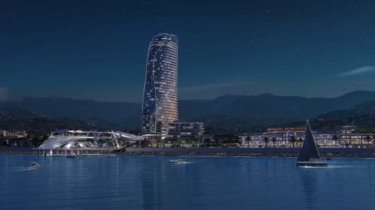 A tower by the water in nighttime Batumi, Georgia, with a mountain backdrop and boats against a starry sky.