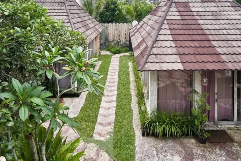 Tropische Bungalows mit einem Garten und einem Weg aus grünem Gras, umgeben von blühenden Pflanzen.