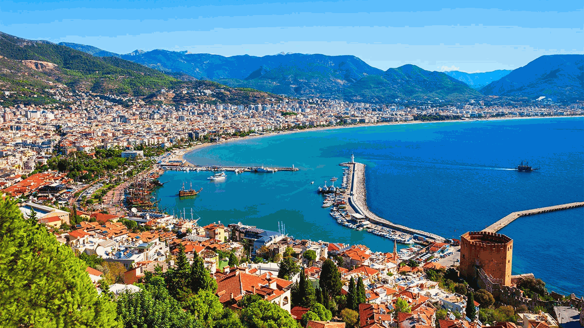 Panorama of the coast with the harbor and the red tower in the Turkish city of Alanya, surrounded by mountains and green hills.