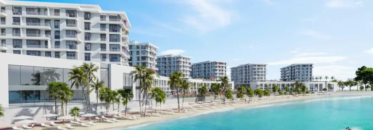 Modern buildings by the shoreline with palm trees and lounge chairs on the beach under blue skies.